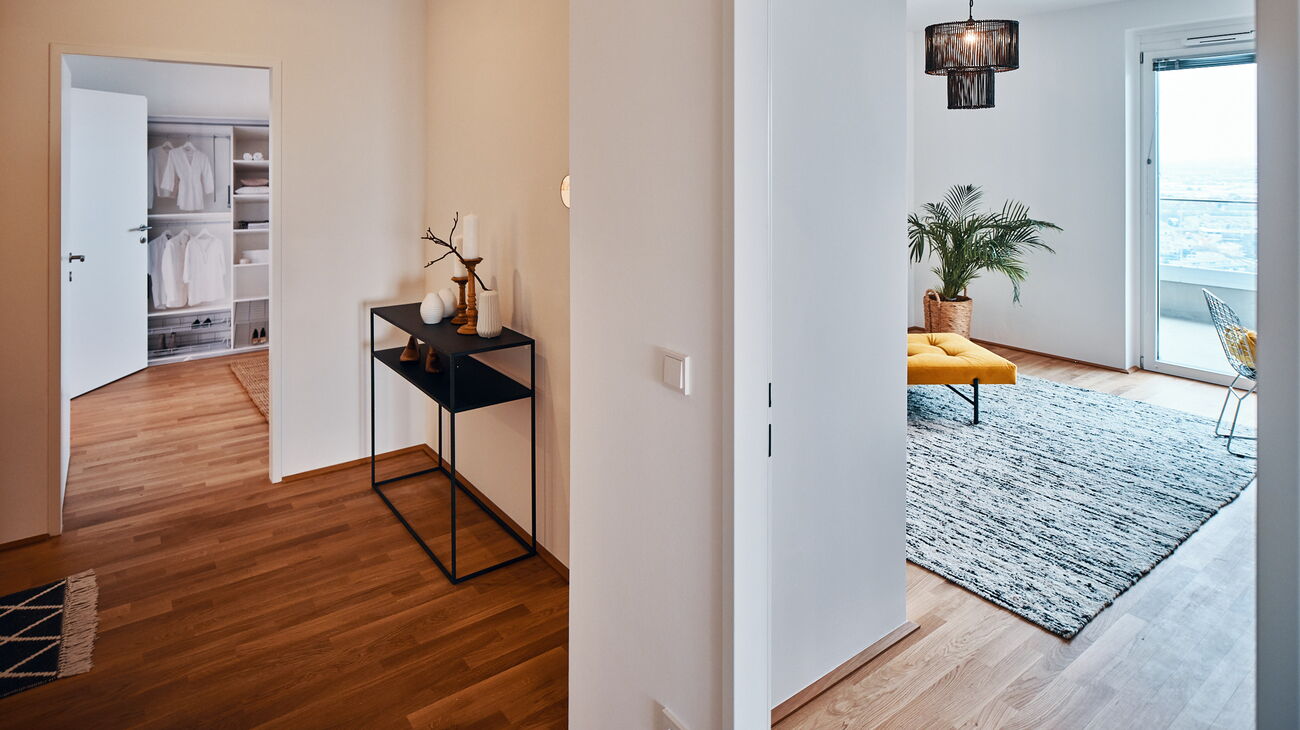 Eingang in L-Form mit Holzboden und schwarzer Metallkonsole und Blick ins Schlafzimmer sowie in ein Arbeitszimmer mit senfgelbem Daybed, schwarzer Rattanleuchte und weiß-schwarzem Wollteppich und einer Palme im Seegras-Übertopf in der Ecke