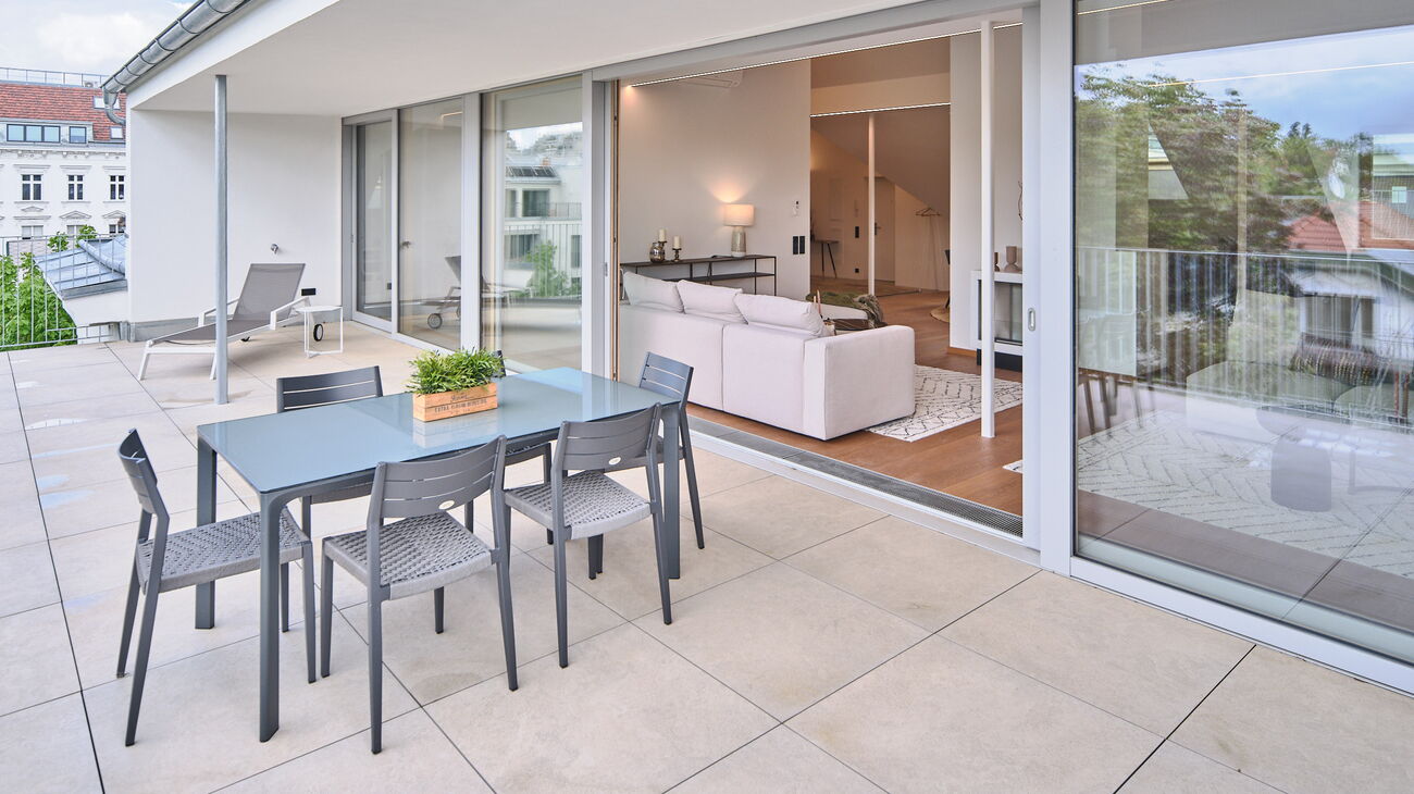 Terrasse mit Steinfliesen und grauem Esstisch samt grauen Stühlen - Blick ins Wohnzimmer