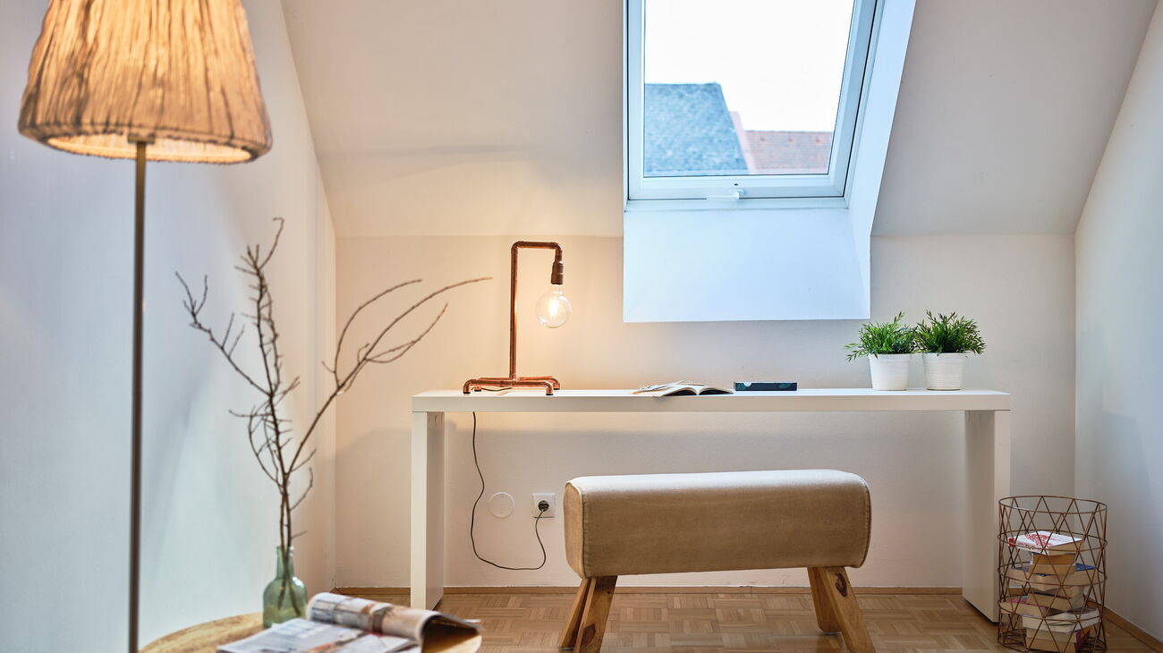 Mini-HomeOffice: Blick vom Sessel auf Stehleuchte mit Stoffschirm, Beistelltischchen aus Sperrholz, weiße Hochglanz-Konsole mit Sitzbank wie eine alte Turnbank, Tischleuchte aus Kupferrohren mit Glühbirne, Gitterkorb Kupfer mit Büchern, grüne Glasvase