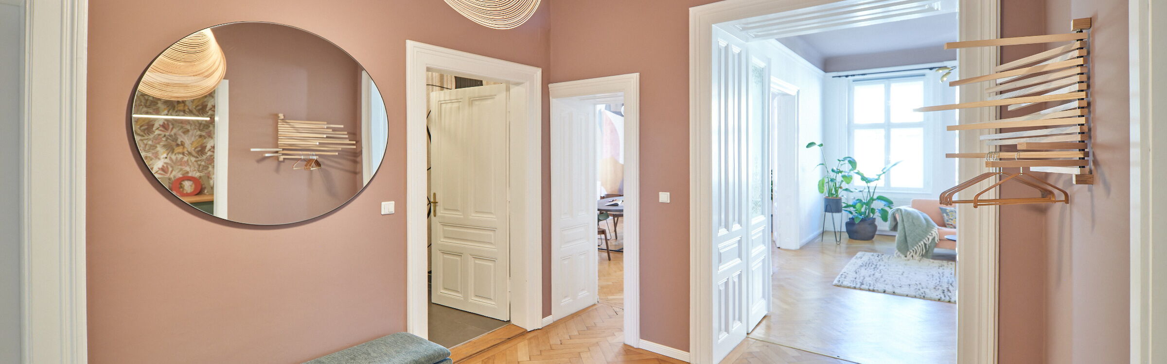 mauvefarbenes Vorzimmer links mit rundem Spiegel und rechts mit Holz-Wandgarderobe, Parkttboden und Sisalteppich und ganz feiner Holzfurnier-Design-Deckenleuchte