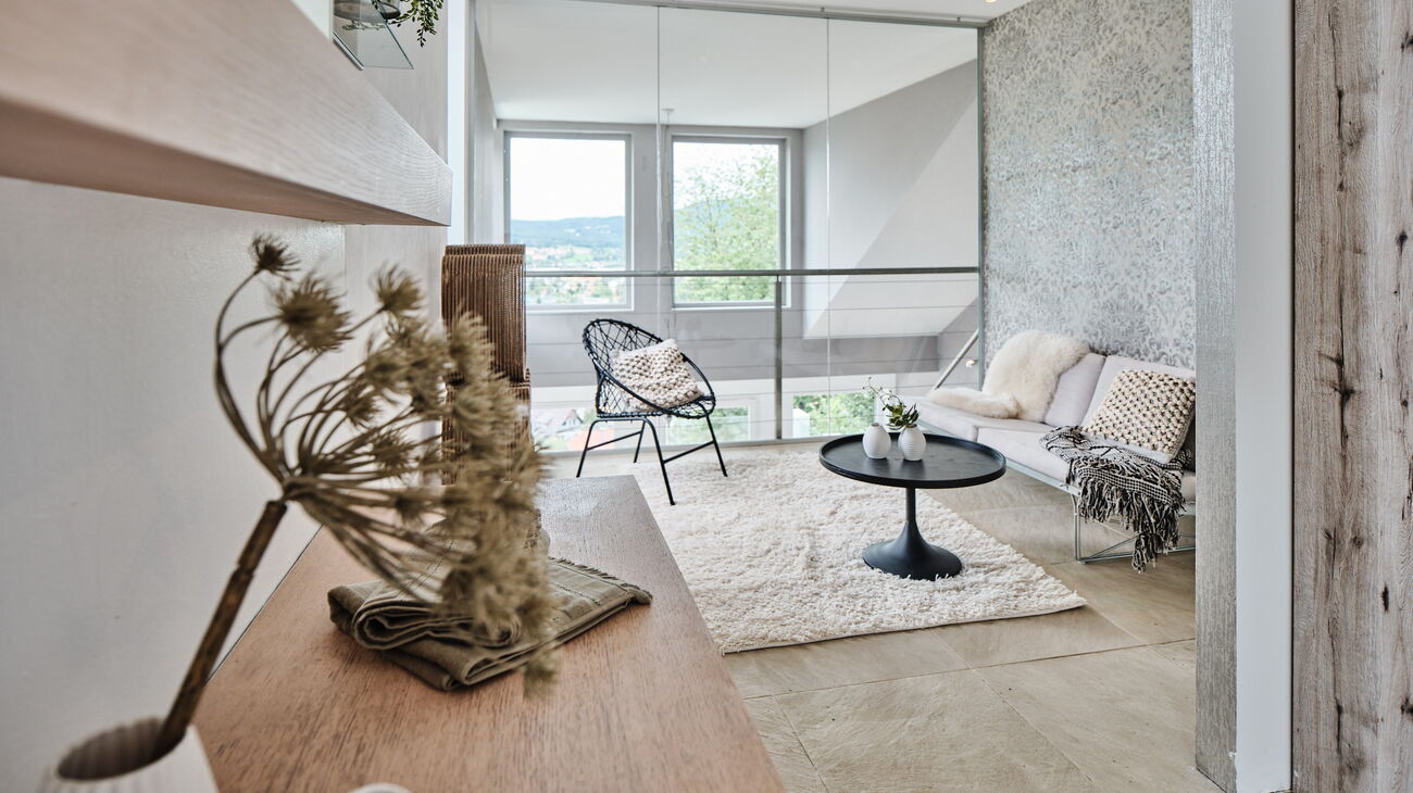 Galerie symbolisiert als Gästezimmer mit Sofa in beige, rundem Couchtisch in schwarz und Kuschelteppich in creme, dazu Holzregale mit Vasen und Blüten und Stehleuchte mit Kugeln in creme