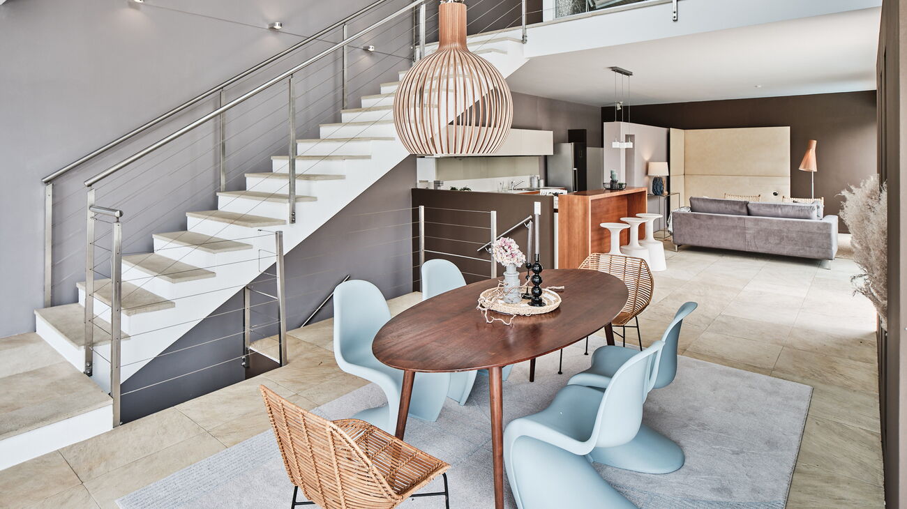 Essplatz neben der Stiege mit grauer Wand, ovaler Holztisch und verschiedene Stühle in Rattan, Pantone Chairs in eisblau und eine Secto Leuchte aus Holz mit weichem Teppich in graublau