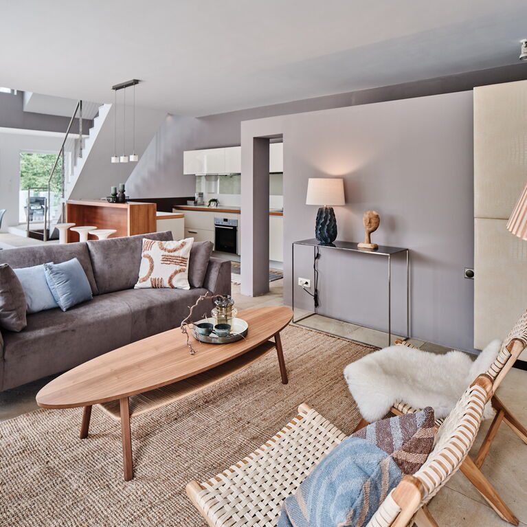 Offener Wohnraum mit grauem Samtsofa ovalem Couchtisch in Holz Loungestühlen in Holz weißer Küche mit Bar aus Holz und dahinter ein Essplatz mit Holztisch in oval und einer Secto Leuchte aus Holz sowie modernen Kunststoffstühlen Wandfarbe grau und braun