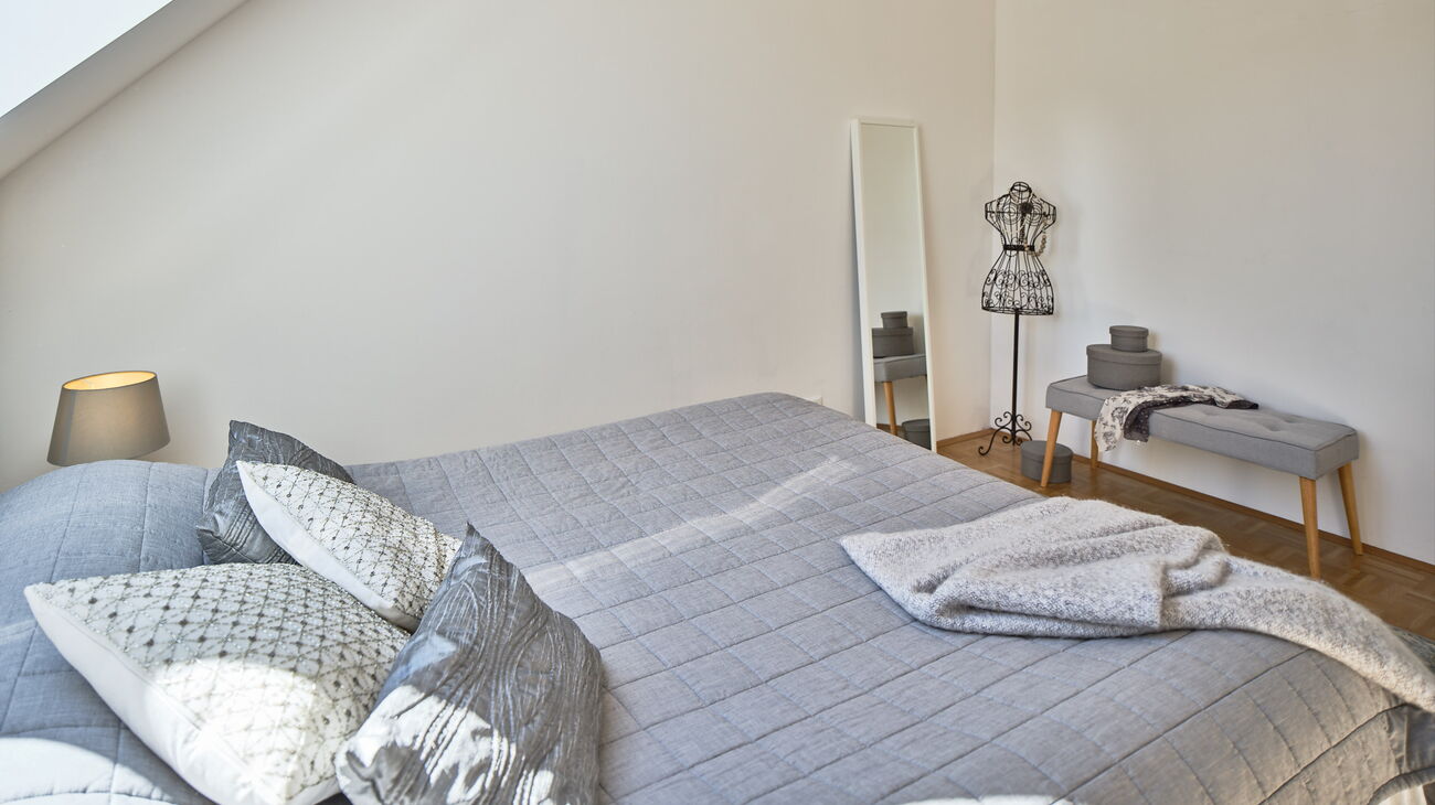 Bett mit grauer Bettdecke und unterschiedlichen Zierkissen in Grautönen und Strickplaid, graue Tischlampe, Sitzbank grau mit Holzbeinen und Dekoschachteln, Standspiegel weiss, Kleiderpuppe aus Metall