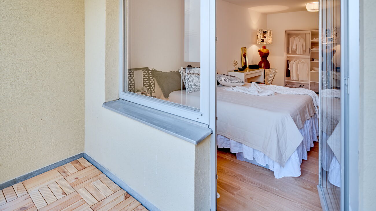 Blick vom Balkon ins Schlafzimmer mit Boxspringbett, weißer Schminktisch mit Metallstuhl, Stehleuchte aus alter Holz-Schneiderpuppe