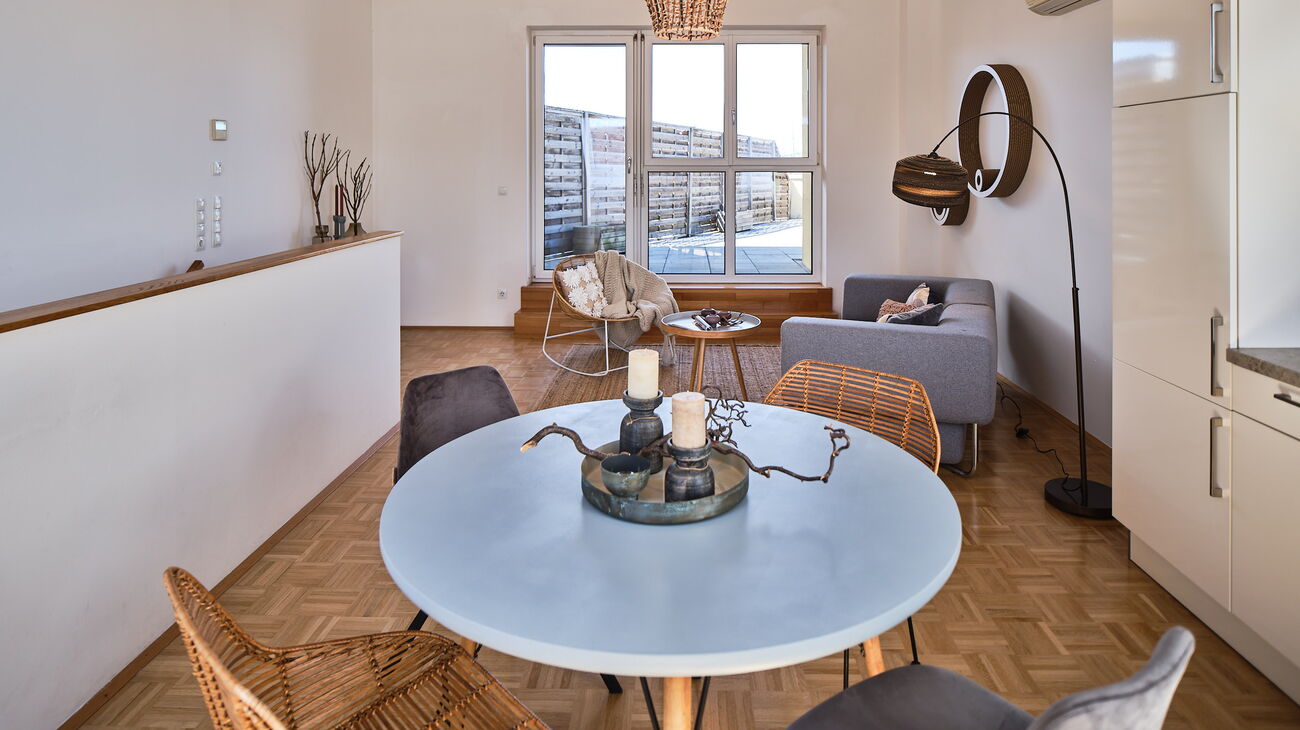 Wohnzimmer mit Essbereich: graues Stoffsofa mit Augestell 2teilig, Sisalteppich, Loungestuhl aus Rattan, kleiner Beistelltisch aus Holz, Bogenleuchte mit Kartonschirm, Wanddeko aus Kartonkreisen, runder Esstisch Holz mit 4 unterschiedlichen Stühlen