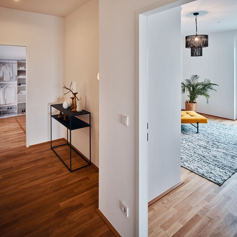 Vorzimmer mit schwarzer Konsole und Blick ins Schlafzimmer sowie in ein Arbeitszimmer mit Schreibtisch in Holz/schwarz und senfgelbem Daybed und schwarz weißen Wollteppich sowie schwarzer Rattan-Deckenleuchte und einer Palme in Seegras-Übertopf