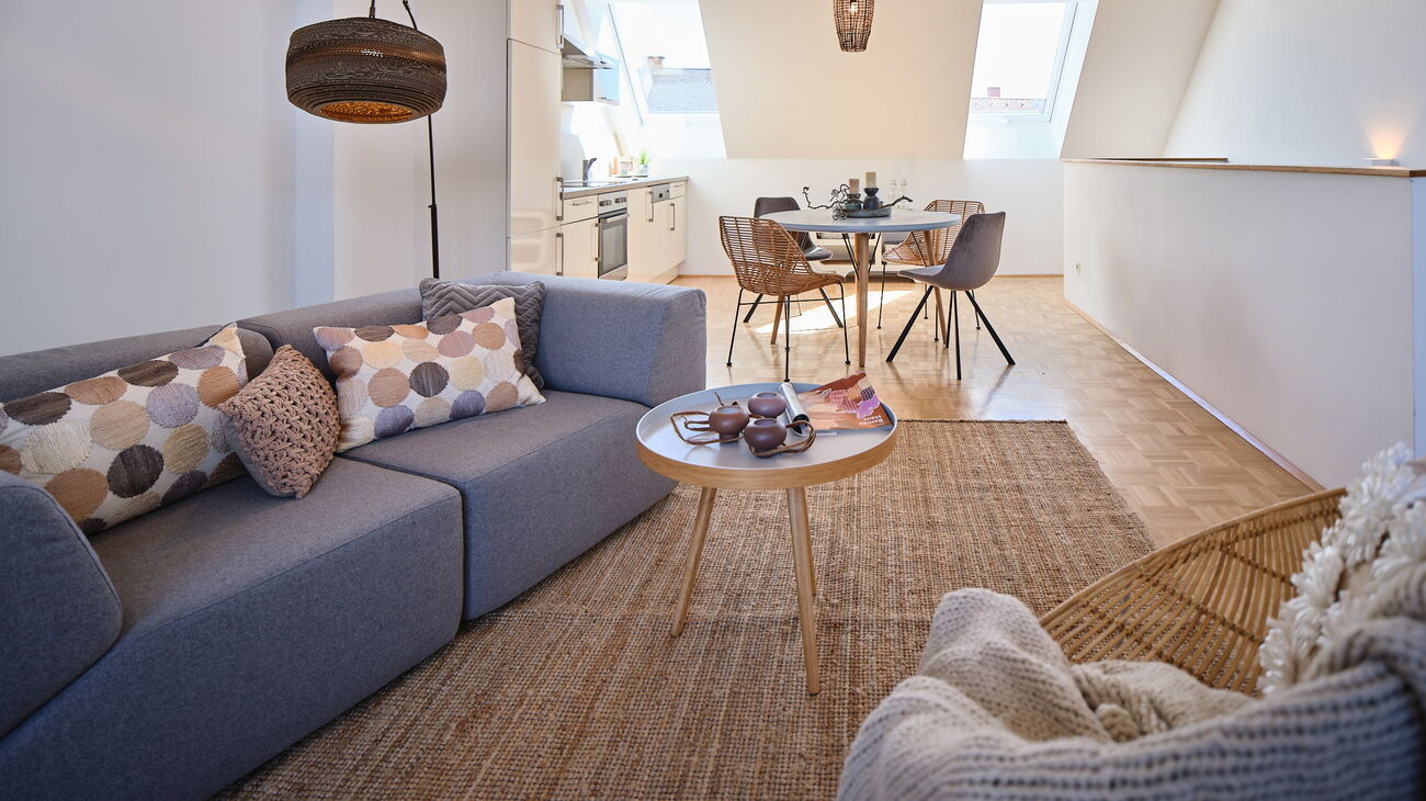 Wohnzimmer mit Essbereich: graues Stoffsofa mit Augestell 2teilig, Sisalteppich, Loungestuhl aus Rattan, kleiner Beistelltisch aus Holz, Bogenleuchte mit Kartonschirm, Wanddeko aus Kartonkreisen, runder Esstisch Holz mit 4 unterschiedlichen Stühlen