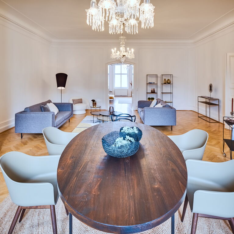 Großer Wohnraum mit Erkerzimmer, Kristalllustern, im Erker ein Essplatz mit massivem Holztisch und modernen olivgrünen Stühlen, dahinter graue Sofas und Loungechair in beige und Couchtisch-Set in Metall