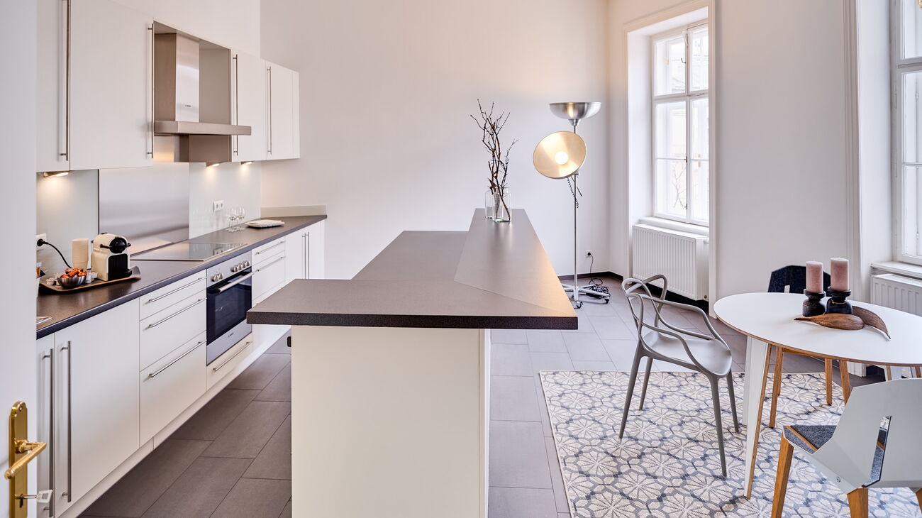 Wohnküche weiß mit schwarzer Arbeitsplatte, rundem Tisch in weiß mit 4 Stühlen in grau, Teppich mit Blumenmuster und große Stehleuchte mit 2 halbrunden silbernen Schalen