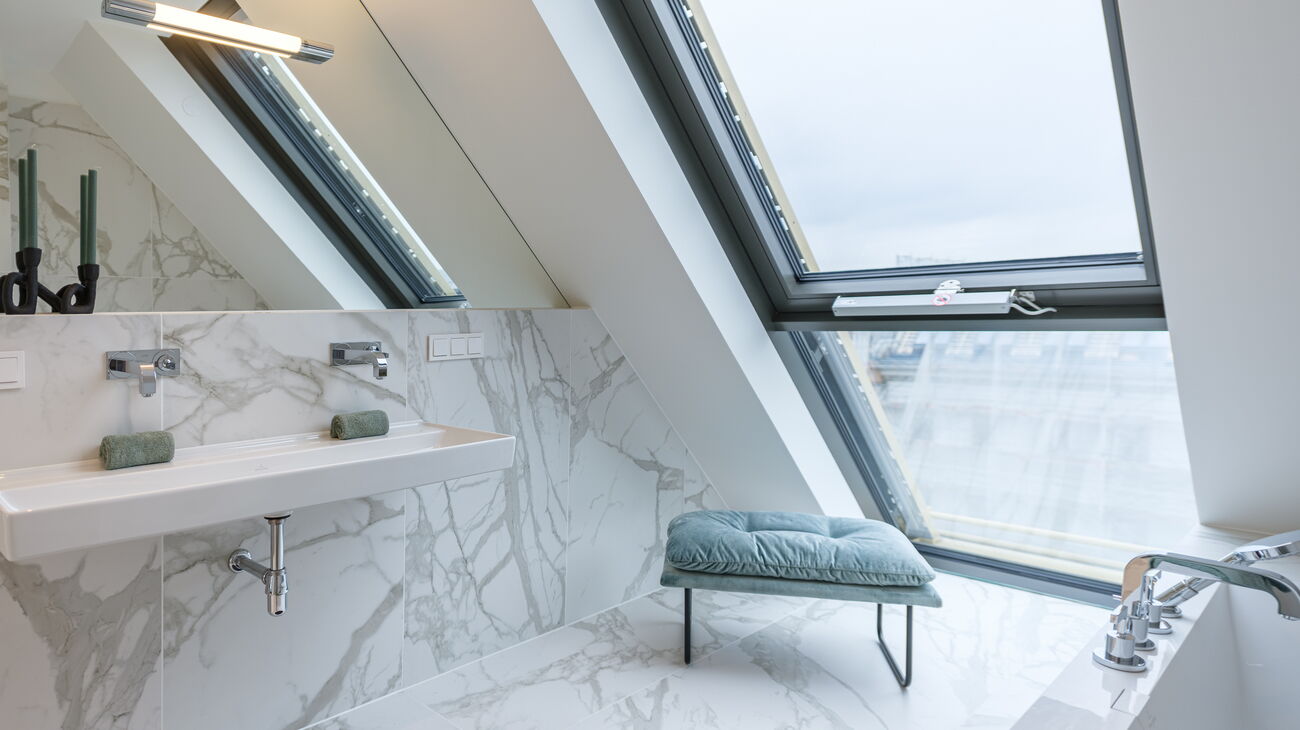 Marmorbad in weiß-grau mit weißem Waschbecken und schwarzen Kerzenleuchtern vor dem eingelassenen Wandspiegel, mintfarbene Sitzbank mit schwarzen Metallbeinen, schwarzer runder Bad-Teppich und mintgrüne Handtücher auf der Badewanne vis a vis
