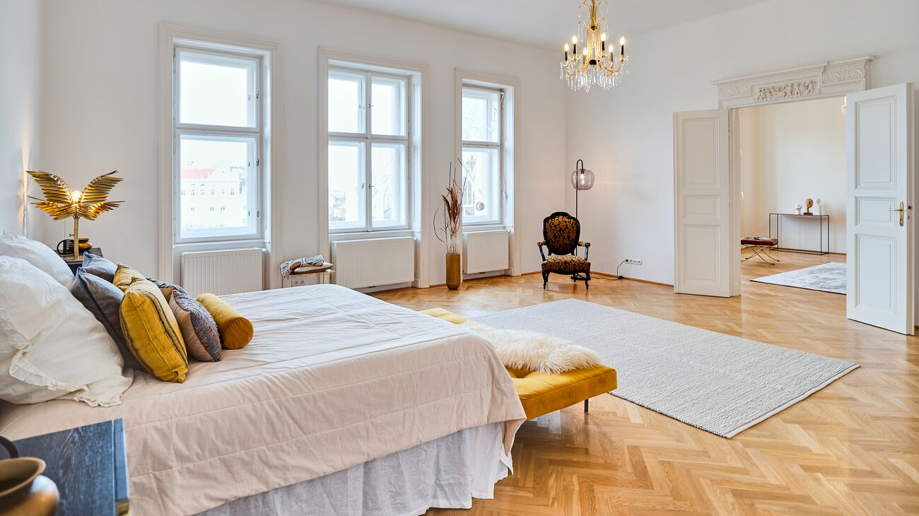 Schlafzimmer mit Boxspringbett, Wollteppich, schwarzen Nachttischen, altem Lesesessel in blau-gelb, gelber Sitzbank und Stehleuchte aus Glas sowie traditionellem Luster