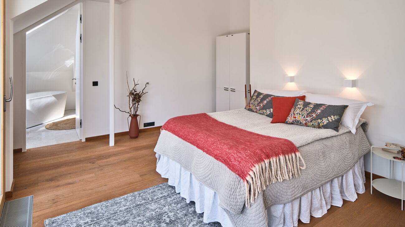 Schlafzimmer Bett mit grauer Bettdecke und rot-grauen Accessoires, grauer Nachttisch, rote Bodenvase und grauem Teppich sowie Wandleuchten in Würfelform