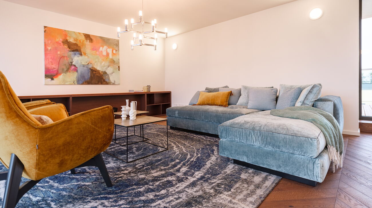 großer Wohnraum mit mintgrünem Samtsofa goldig-schwarzem Couchtisch-Set großen gelben Samt-Loungestühlen Vintageteppich in blau mit Kreisen sowie Design-Chrom-Luster 12-armig mit matten Glaszylindern und abstaktem Bild in kräftigem Pink-Gelb-und Braunton