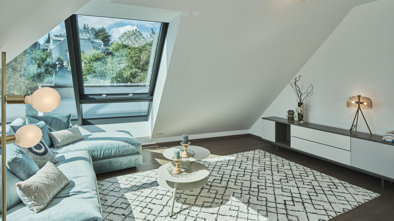 Dachgeschoß-Wohnraum mit 2 Deckenspots, goldene Stehleuchte mit Bulbs im Vordergrund, L-förmiges Samtsofa in mintgrün, Teppich Baumwolle creme mit schwarzen Rauten, 3-Ebenen-Couchtisch Metall in taupe, Lowboard in weiß/grau mit Laden, Tischleuchte gold