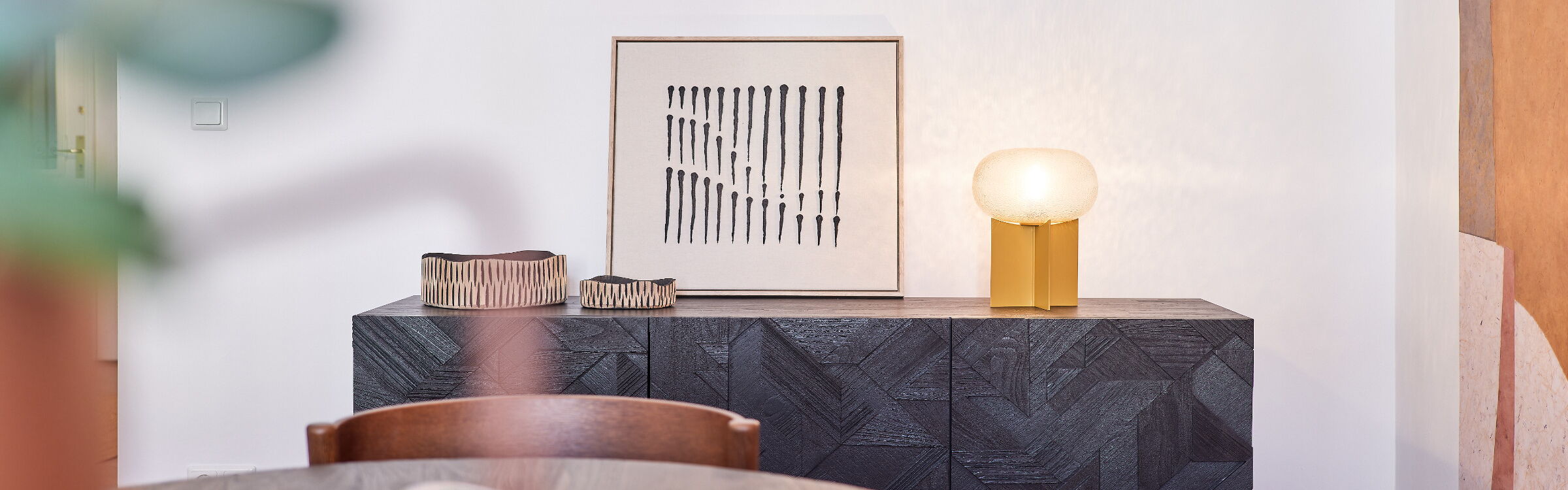 schwarzes Sideboard mit geschnitzter Front darauf senfgelbe Tischleuchte und ein modernes Bild schwarz auf beigem Leinen sowie 2 Holzschalen in creme-schwarz daneben