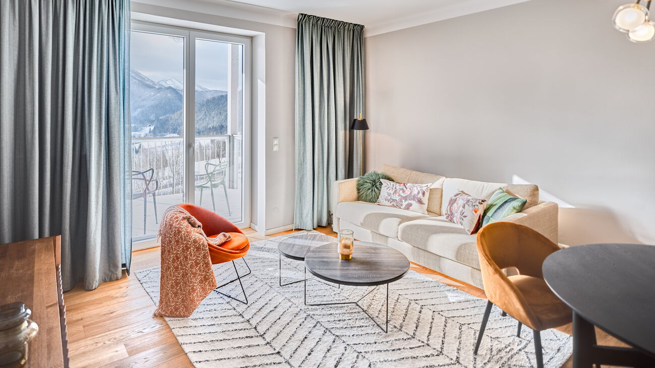 Wohnzimmer mit beigem Schlafsofa, orangem Samt-Loungestuhl und beigem Woll-Teppich