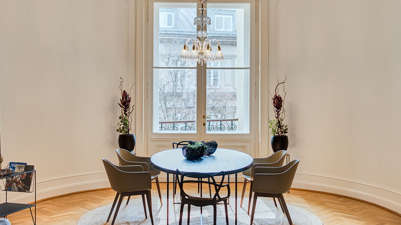 Wohnzimmer mit Luster, grauem Sofa, Couchtisch-Set aus Metall mit Holzplatte, Wollteppich in grau-gelb-Tönen, und im runden Erker massiver Esstisch aus Holz mit schwarzem Metallgestell, Design-Stühle mit Holzbeinen und salbeifarbenen Kunststoffschalen