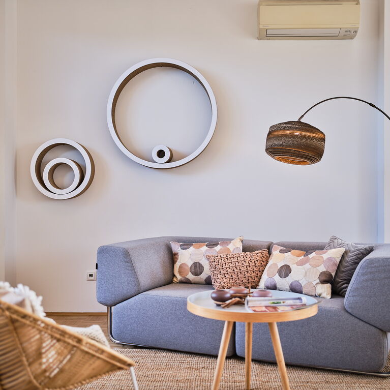 Designsofa grauer Stoff 2teilig mit Alugestell, Bogenleuchte mit Kartonschirm, Wanddeko aus Kartonkreisen, Rattanstuhl, Sisalteppich, kleiner Holzbeistelltisch