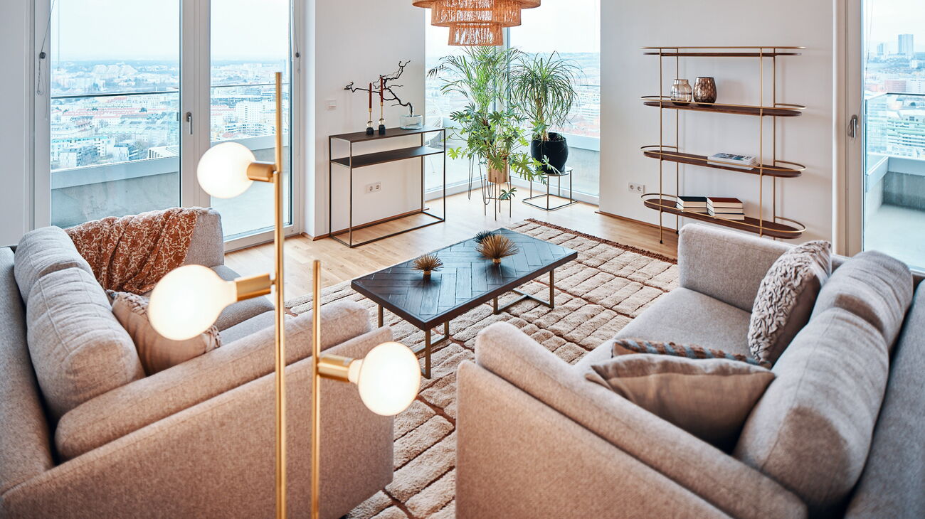 offener Wohnraum mit Blick über Wien, 2 beige Stoffsofas, Holzcouchtisch, Bücherregal in ovaler Form mit Holz und Metall, Teppich aus beiger Wolle, schwarz Konsole und ein paar Grünpflanzen am Fenster in Metallübertöpfen auf Beinen und Bast-Hängeleuchte