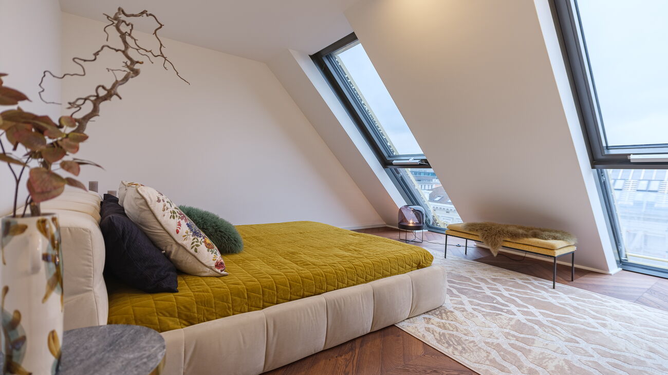 Doppelbett mit wuchtigem Stoffbezug in creme und senfgelber Überdecke mit bunten Kissen, davor Sidetable in Marmor und Vase mit Zweigen, dahinter senfgelbe Sitzbank auf cremefarbenem Teppich und Bodenleuchte mit halbrundem getöntem Glasschirm und Messing
