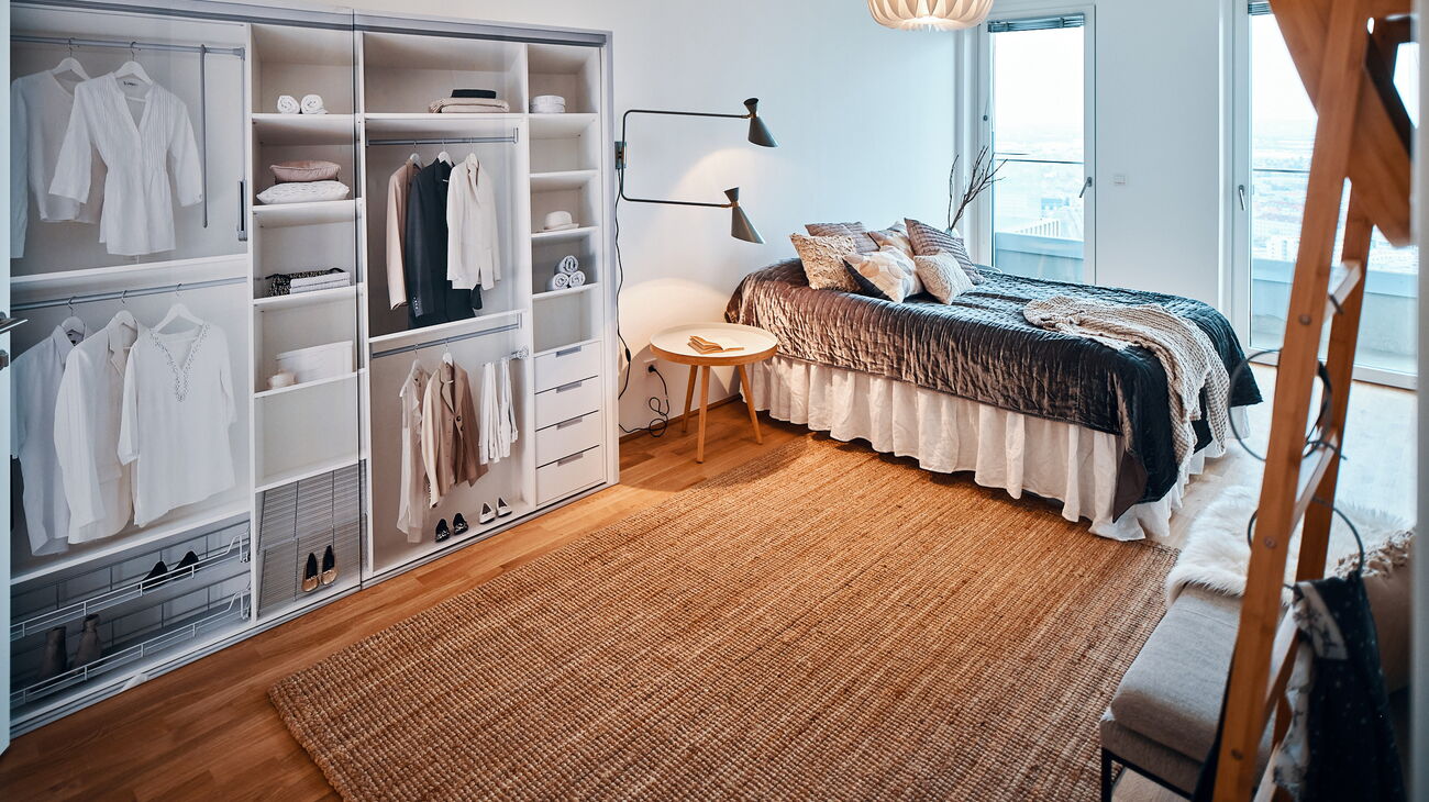 Schlafzimmer mit Dummy-Schrankwand, Doppelbett mit grauem glänzendem Überwurf, weißer Papierfaltenleuchte, Wandleuchte zweiarmig grün als Leselicht und kleinem Beistelltisch aus Holz, Sitzbank und Stehgarderobe wie Holzleiter und Sisalteppich