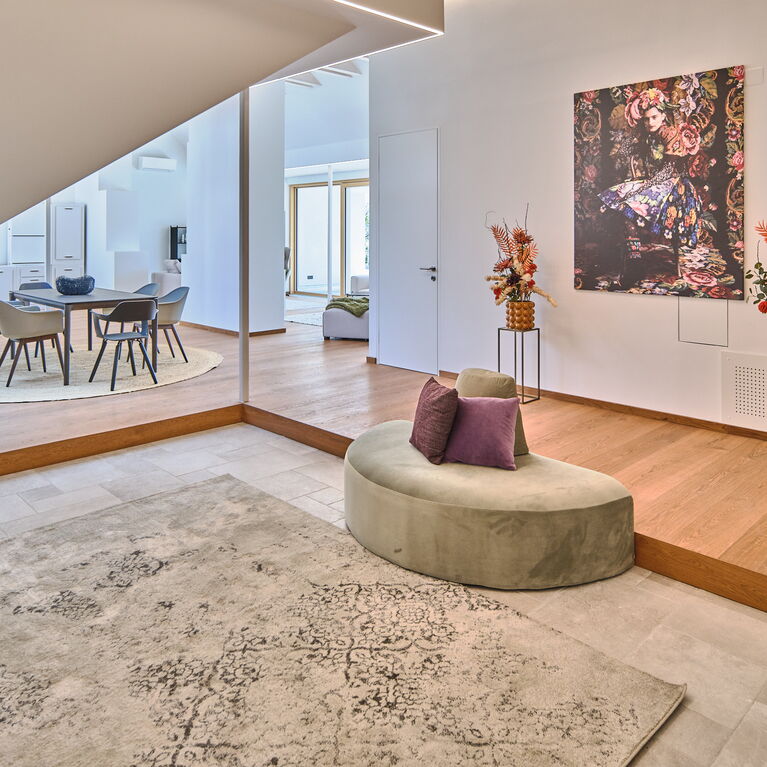 offener Eingangsbereich mit Parkett, silbrigem Teppich, halbrundem mintfarbenen Sofa, großem Bild von Susanne Bisovsky und Gestecken in gelben Töpfen auf schwarzen Pflanzständern