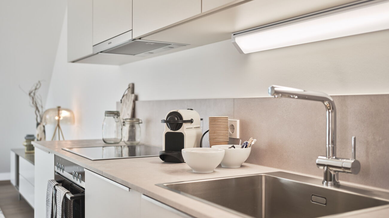 Einbauküche in weiß mit grauer Platte inkl Oberschränken, Nespressomaschine und Becher, Geschirrtücher an der Herdstange, Blick ins Wohnzimmer Richtung Lowboard