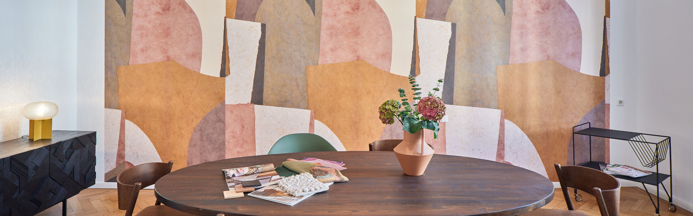 Altbauraum mit massivem ovalem Teak-Tisch und 6 Stühlen vor einer großgemusterten Retro-Tapete in rosa-gelb-grün-braun-beige-Tönen, rechts ein schwarzer Metallservierwagen und links ein schwarzes Sideboard mit geschnitzter Oberfläche