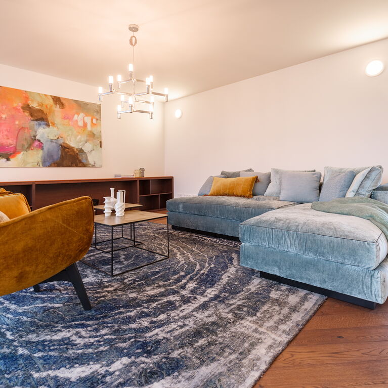 großer Wohnraum mit mintgrünem Samt-Ecksofa goldigem Metall-Couchtisch-Set eckig gelben großen Samt-Loungestühlen dunklem Holz-Lowboard Design-Chromluster und blauem Vintageteppich sowie abstraktem Bild in kräftigen Farben