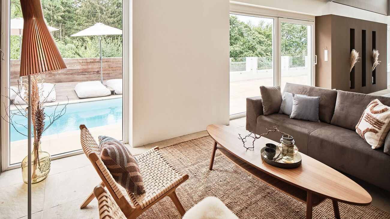 offener Wohnraum mit Blick auf die Pool-Terrasse, graues Samtsofa, Secto Stehleuchte aus Holzlamellen, Loungestühle aus Holz, ovaler Couchtisch aus Holz und Sisalteppich