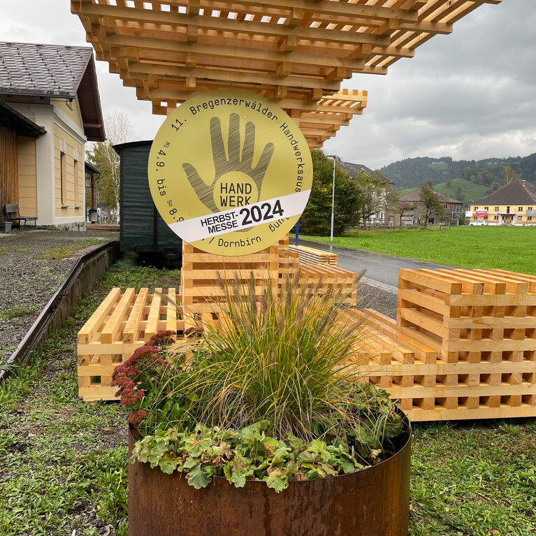 Holz-Konstrukt am Eingang zum Werkraumhaus im Bregenzerwald mit rundem Metallschild HANDWERK+FORM und grossem runden Blumentopf grün bewachsen davor